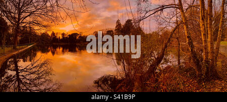 Brynmill Park a Swansea, Regno Unito al tramonto. Cielo arancione con alberi riflessi sulla superficie dell'acqua. Foto Stock