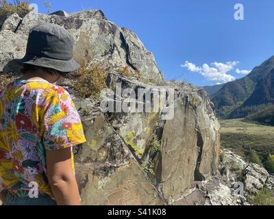 Una ragazza viaggiante in un cappello e una T-shirt con fiori si avvicina ai dipinti rupestri di un antico uomo sulle montagne Altai. Foto Stock