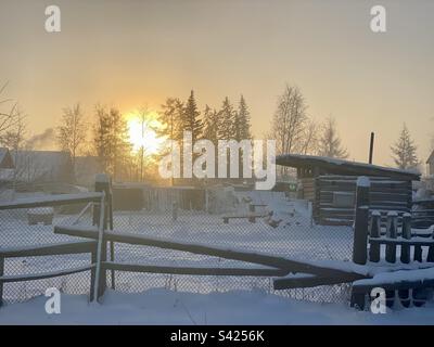 Tramonto in una giornata gelida in inverno dietro gli alberi recinzione e capannoni di cottage privati nel villaggio di Yakutia. Foto Stock