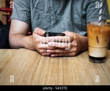 Parte centrale dell'uomo che usa il telefono cellulare a tavola con una bevanda ghiacciata a base di tè al latte. Foto Stock