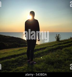 Una donna in silhouette al tramonto che cammina sopra il Mare d'Irlanda su una sezione drammatica del popolare percorso della Costa Ceredigion vicino a Llangrannog nel Galles occidentale Foto Stock