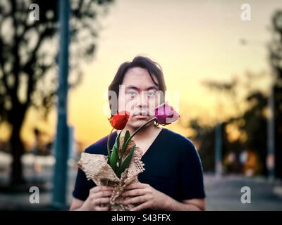 Giovane asiatico che tiene due tulipani, di colore rosso e viola, contro gli alberi, lampione e cielo tramonto. Foto Stock