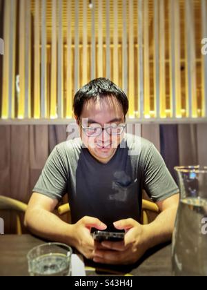 Sorridente giovane asiatico in occhiali con smartphone al tavolo all'interno del ristorante. Foto Stock