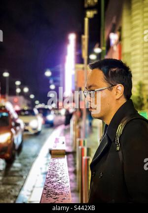 Ritratto di giovane asiatico in attesa sul marciapiede contro le luci al neon, gli edifici e la strada trafficata nella città di notte. Messa a fuoco selettiva. Colorato. Foto Stock