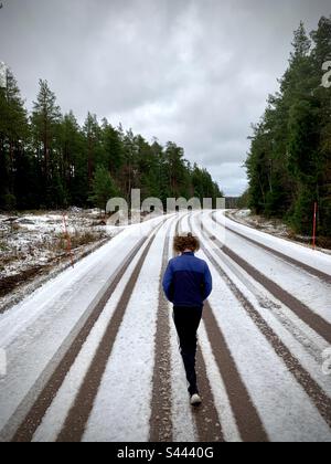 Un giovane adolescente che cammina da solo nel profondo del pensiero su una strada invernale coperta di neve in neve con le piste per pneumatici auto Foto Stock