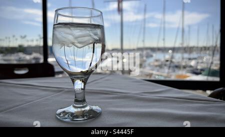 Bicchiere d'acqua ghiacciata con alberi di barca concentrati attraverso di essa, e soft focus sugli yacht ormeggiati a Marina del Rey, California, sullo sfondo Foto Stock