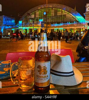 Bottiglia di birra Singgah e cappello da viaggio che guarda alla stazione ferroviaria di Hua Lamphong a Bangkok in Thailandia di notte Foto Stock