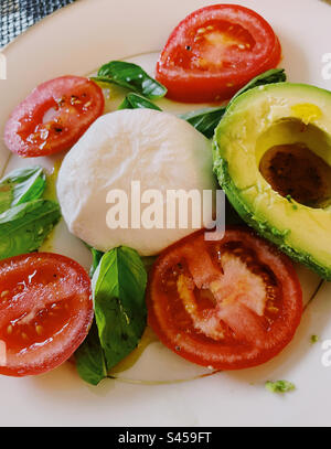 Primo piano di un'insalata tricolore italiana di formaggio Burrata, basilico fresco, pomodori stagionati con vite e mezzo avocado, 2023, USA Foto Stock