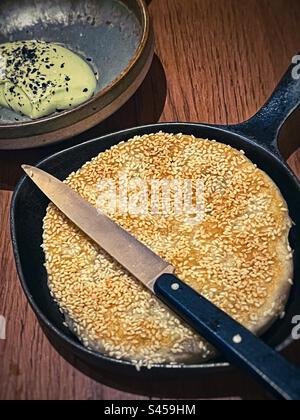 Vista angolare della focaccia di sesamo in padella con coltello e avocado cremoso con semi di sesamo neri immersi nella ciotola sul tavolo. Foto Stock