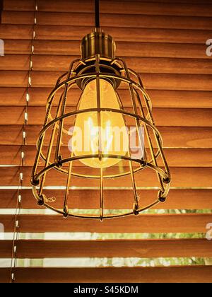 Primo piano della lampadina decorativa in stile Edison vintage che si illumina di una luce gialla soffusa in una gabbia metallica retrò contro le veneziane. Stile vintage. Retro. Design degli interni. Foto Stock