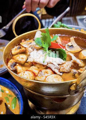 Vista dall'alto della piccante zuppa thailandese Tom Yum con funghi, pollo ed erbe in pentola con una mano che tiene un cucchiaio sullo sfondo. Foto Stock