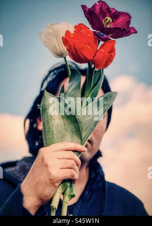Primo piano di un giovane che regge fiori di tulipano multicolori contro il cielo blu con le nuvole. Faccia oscurata. Temi primaverili. Foto Stock