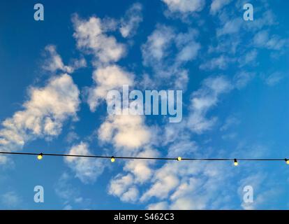 Vista angolare ridotta delle lampadine luminose pendenti dalla linea aerea contro il cielo blu con soffici nuvole bianche con spazio per la copia. Filo di lampadine. Foto Stock