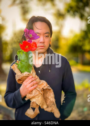 Ritratto di un giovane asiatico che tiene tulipani rossi e viola contro gli alberi e tramonta il sole. Sognante. Concentrati sul primo piano. Ora d'oro. Tema primaverile. Foto Stock