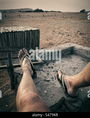 Un uomo con i sandali poggia le gambe su un barbecue mentre si rilassa mentre il sole tramonta in Namibia Foto Stock