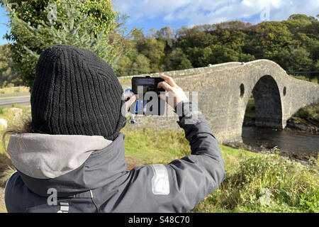 Una femmina con una giacca grigia e un cappello nero che scatta una foto iPhone di un tradizionale ponte in pietra - il ponte di Clachan sull'Atlantico. Foto Stock