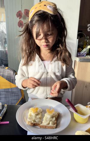 Una bambina asiatica filippina carina che prepara panini con crema di banana su un piatto bianco sul tavolo della cucina. Indossa una maschera gialla per gli occhi dormienti a forma di gattino. Foto Stock