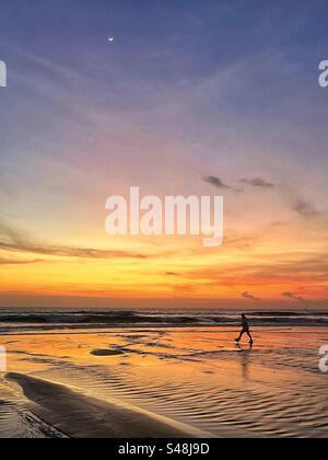 L'uomo che cammina al tramonto sulla spiaggia di Seminyak Bali si staglia contro il mare e il bagliore arancione nel cielo Foto Stock