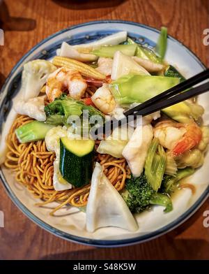 Primo piano ad angolo elevato di spaghetti fritti con frutti di mare e verdure miste su piatto con bacchette sul tavolo. Cibo cinese. Foto Stock