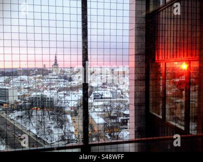 Splendida vista attraverso la finestra con reticolo in rete metallica sui tetti della città invernale di Tallinn con l'alba rossa che si riflette nella finestra successiva Foto Stock