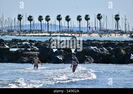 Due ragazzi che giocano tra le onde sulla spiaggia sullo sfondo del molo roccioso e allineano palme a Palavas, Occitanie, Francia Foto Stock