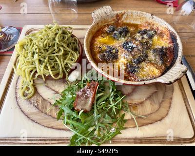 Shot di un piatto di legno del ristorante con un piatto di spaghetti al pesto, polpette in salsa di pomodoro con parmigiano e insalata verde con pancetta. Foto Stock