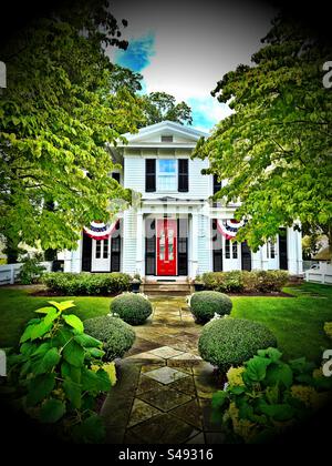 Sentiero che conduce alla casa bianca in legno, con persiane nere e porta rossa. Situato a Essex, Connecticut, Stati Uniti. Molto verde, tra cui alberi, cespugli e altre piante. Foto Stock