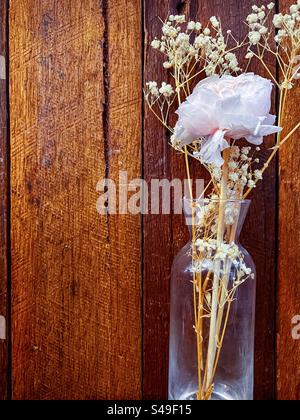 Single pale pink rose and dried white flowers in a glass vase on stained wood background. Cottagecore aesthetics. Vintage style. Still life. Foto Stock