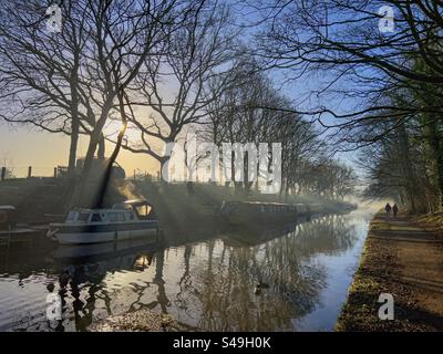 Misty mattina invernale sul canale di Leeds e Liverpool ad Adlington nel Lancashire. Barche strette ormeggiate da una banca e due persone camminano sul sentiero trainato con il sole che sbircia da dietro l'albero. Foto Stock