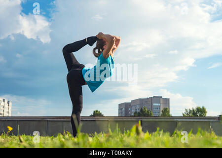 Ragazza sportiva. Giovane bella ginnasta in piedi con il piede sopra la sua testa in uno dei yoga pone nel parco della città sull'erba. Foto Stock