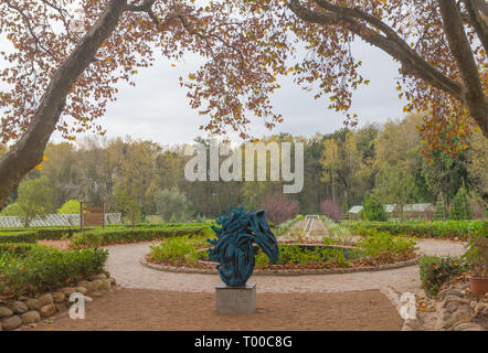 Vista sul giardino in autunno incorniciato da foglie su un albero e con una scultura di un cavallo in centro prima di un percorso circolare attorno ad un isola di piante Foto Stock