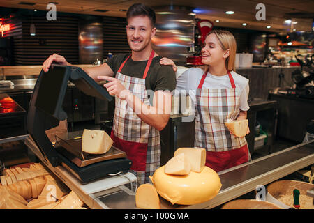 Giovane uomo e donna stand al ripiano di formaggio in un negozio di alimentari. Egli peso formaggio. Modello femminile povera a guy guarda a lui e sorriso. Il paradiso del formaggio. Foto Stock