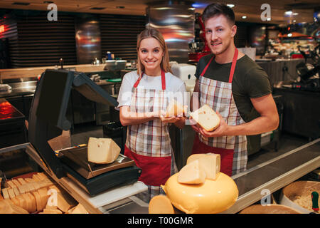 Giovane uomo e donna stand al ripiano di formaggio in un negozio di alimentari. Le persone in attesa di pezzi e pongono sulla fotocamera. Aspetto e sorriso. Foto Stock