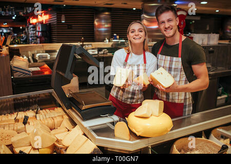 Giovane uomo e donna stand al ripiano di formaggio in un negozio di alimentari. Felice lavoratori positivi pongono sulla fotocamera e il sorriso. Il paradiso del formaggio. Foto Stock