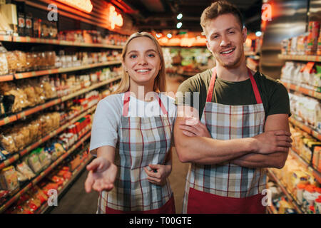 Felice allegro giovane uomo e donna stanno nel negozio di alimentari tra scaffali ad pasta. Essi pongono sulla fotocamera e il sorriso. Giovane donna di raggiungere le mani per telecamera. Foto Stock