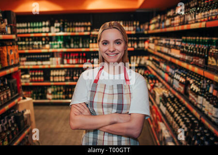 Giovane donna stand in drogheria tra scaffali ad alcool. Ella tenere mani incrociate e sorriso. In posa sulla fotocamera. Indossando il grembiule da soli. Foto Stock