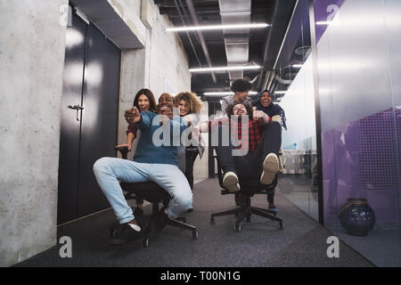 Multiethnics startup business team di sviluppatori di software per divertirsi durante la corsa su sedie da ufficio,entusiasti diversi dipendenti ridere godendo di un divertente Foto Stock