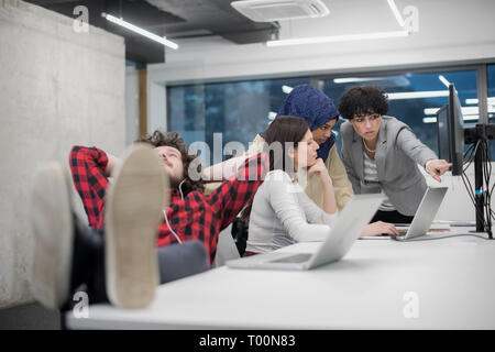 I giovani lo sviluppatore di software in appoggio con le gambe sulla scrivania mentre il suo multiethnics team aziendale la scrittura di codice di programmazione in background all'avvio di office Foto Stock