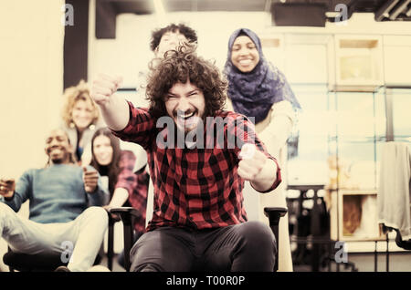 Multiethnics startup business team di sviluppatori di software per divertirsi durante la corsa su sedie da ufficio,entusiasti diversi dipendenti ridere godendo di un divertente Foto Stock