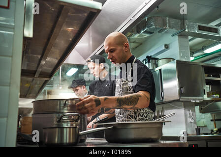 Team di Professionisti. Lo chef concentrato in grembiule, con tatuaggi sulle sue braccia Cucinare con i suoi due assistenti in un ristorante di cucina. Concetto di cucina a vista Foto Stock