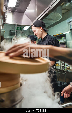 Il processo di cottura. Stretta di mano dello chef holding coperchio di legno durante la preparazione di alimenti con i suoi due assistenti in un ristorante di cucina. Foto verticale. Concetto culinario Foto Stock