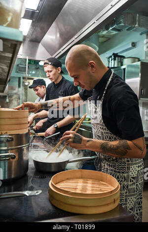 La massima concentrazione. Giovani chef calvo in grembiule con tatuaggi sulle sue braccia preparare il piatto con i suoi due assistenti in un ristorante di cucina. Il processo di cottura Foto Stock