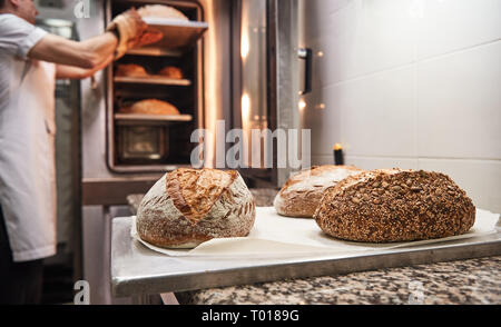 Famosa panetteria. Chiusura del pane appena sfornato sul bordo con baker tenuto caldo il pane dal forno sullo sfondo. Concetto di panetteria. Foto Stock