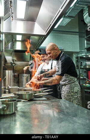 Molto caldo allegro chef e due suoi assistenti preparano il piatto sul fornello con un fuoco aperto nella cucina del ristorante. Flambe. Concetto di cucina a vista Foto Stock