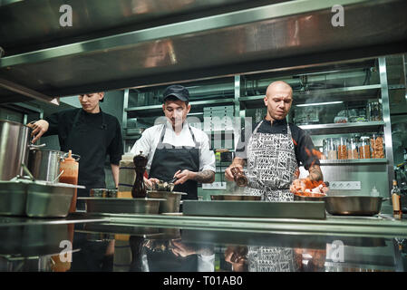 Team di Professionisti. Giovani chef brutale in flambeing uniforme in una cucina professionale. Flambe cibo Foto Stock