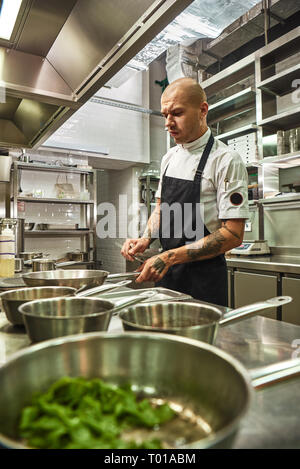 Ristorante Cucina. Foto verticale di concentrato di chef in grembiule con parecchi tatuaggi sulle sue braccia tenendo una padella. Concetto di cibo Foto Stock