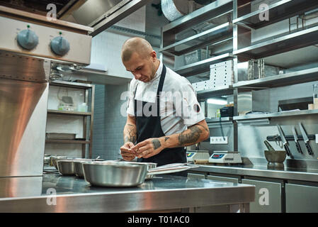 Io amo il mio lavoro concentrato chef maschio con bellissimi tatuaggi sulle sue braccia la scelta di ingredienti per il suo piatto in un ristorante di cucina. Il processo di cottura Foto Stock