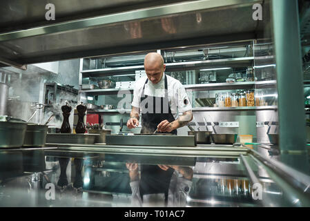 Chef professionale. Famoso giovane chef in grembiule nero a lavorare al suo moderno ristorante cucina. Il processo di cottura Foto Stock