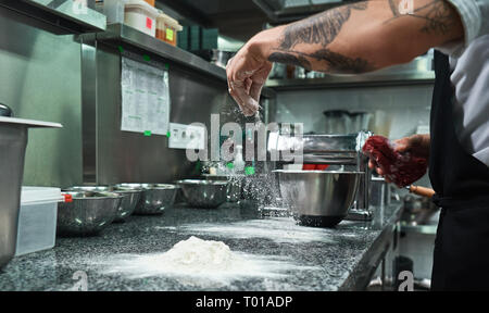 Il processo di cottura. Lo chef maschio le mani con il nero di tatuaggi versando la farina sul tavolo da cucina. Concetto di cibo Foto Stock