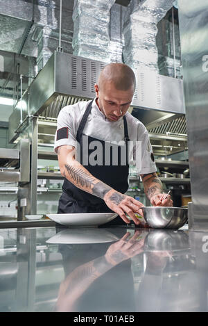 Gli ultimi dettagli. Ritratto verticale di attraente chef maschio con bellissimi tatuaggi sulle sue braccia guarnendo il suo piatto mentre in piedi in un ristorante di cucina. Il processo di cottura. Foto Stock